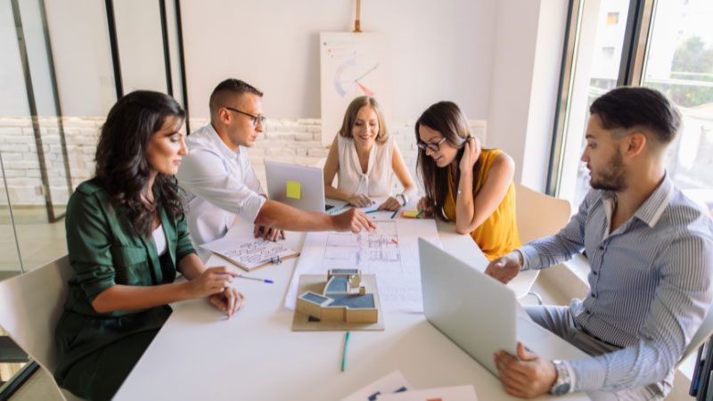 A group of professionals collaborating around a table in a modern office setting, discussing documents and working on laptops. The scene illustrates teamwork and efficiency, key aspects of BPO services (Business Process Outsourcing), where companies delegate tasks to external teams to streamline operations.