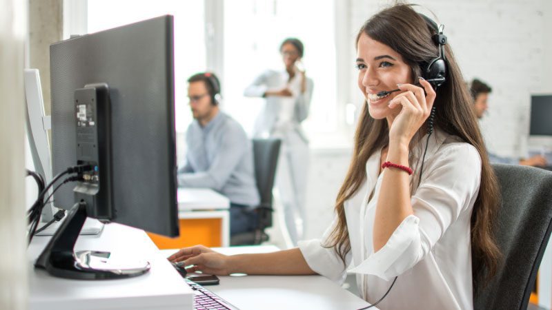 A smiling female representative providing online customer service while wearing a headset and working on a computer in a bright, modern office. Other colleagues in the background are also engaged in customer support, emphasizing the collaborative and efficient nature of online customer service in handling client inquiries.