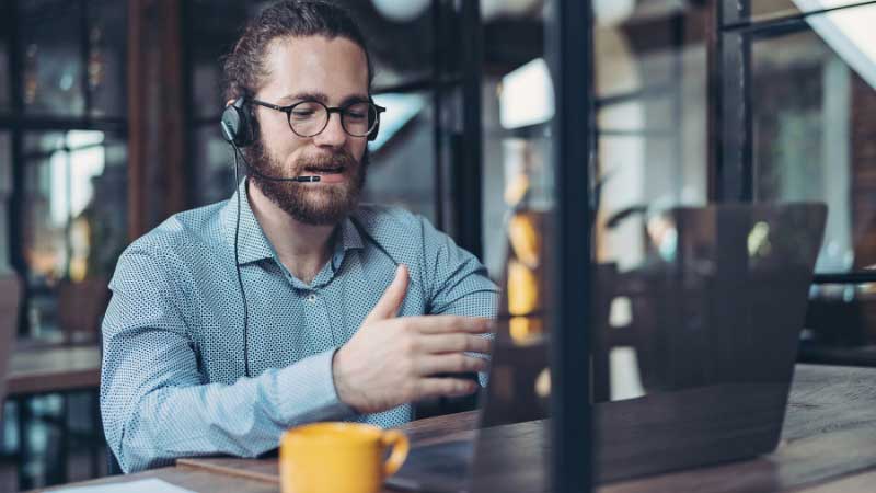 A bearded support agent wearing a headset, speaking to a customer on a video call, offering multilingual customer support solutions.