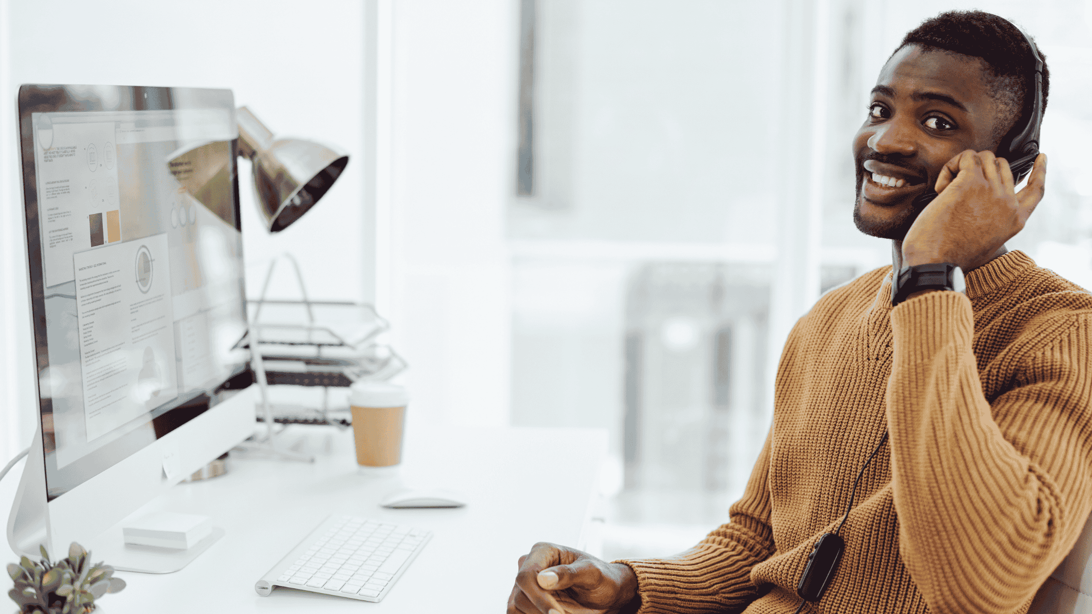 A smiling customer support agent wearing a headset, seated at a desk with a computer, providing multilingual tech support around the clock.