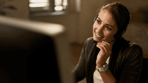 A smiling female support agent wearing a headset and assisting a customer from her workstation