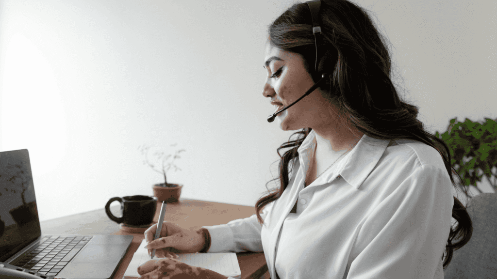 A female customer service agent with a headset, writing notes while working on a laptop at a minimalist desk