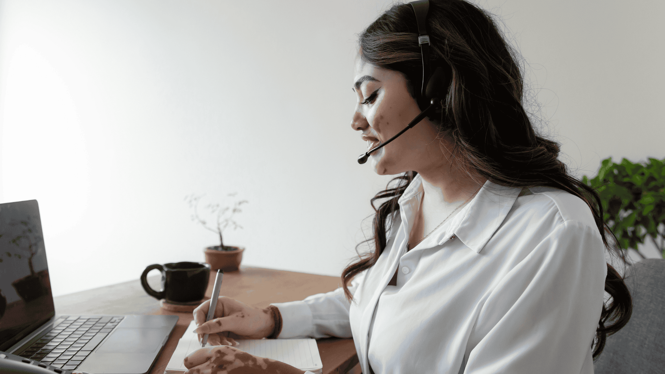 A female customer service agent with a headset, writing notes while working on a laptop at a minimalist desk