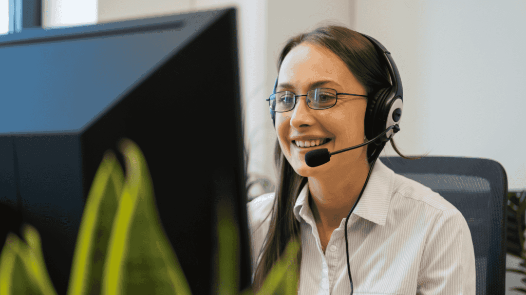 A smiling female call centre agent with glasses and a headset, engaging with a customer on her computer