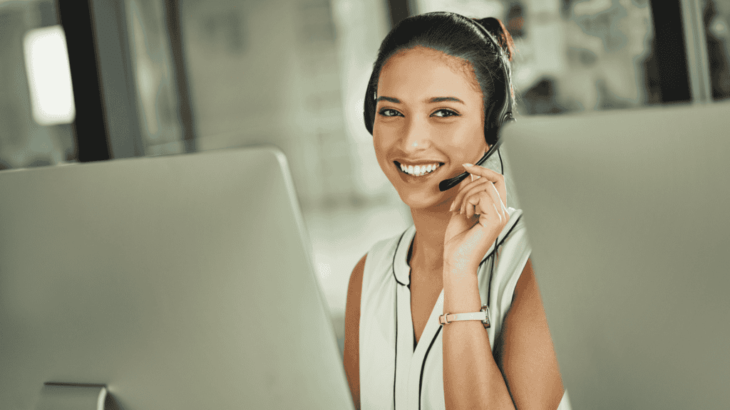 A smiling customer support agent wearing a headset, sitting at a desk and talking to a customer