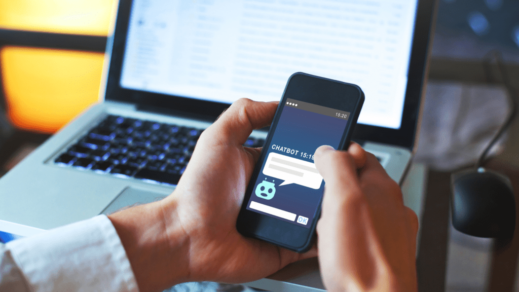 A person interacting with a chatbot interface on their smartphone while working on a laptop in the background