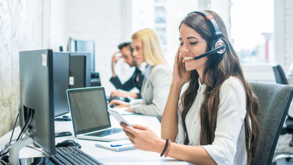 A smiling customer service agent with a headset, assisting customers while working in a modern office alongside her team
