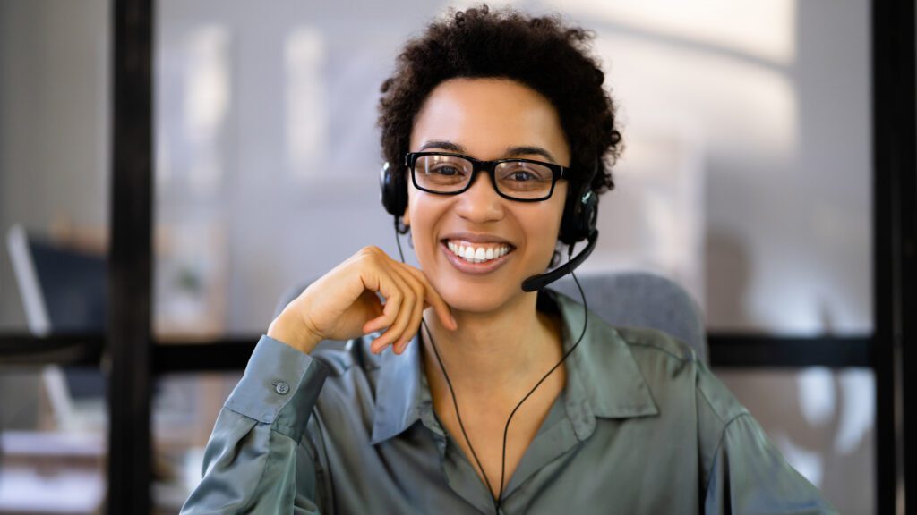 Smiling customer service agent offering multilingual phone support 24/7 in a professional office environment.