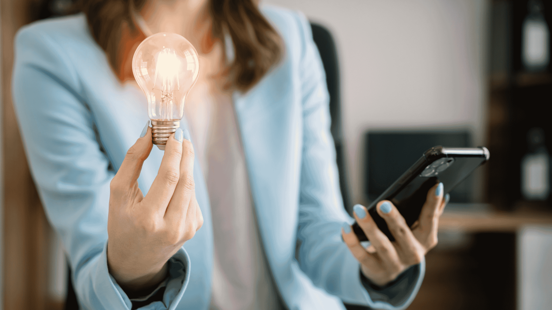 A businesswoman in a light blue blazer holding a glowing light bulb in one hand and a smartphone in the other, symbolizing innovative ideas and technology integration.