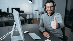 A smiling customer support representative giving a thumbs-up while working on a computer in a professional office setting.