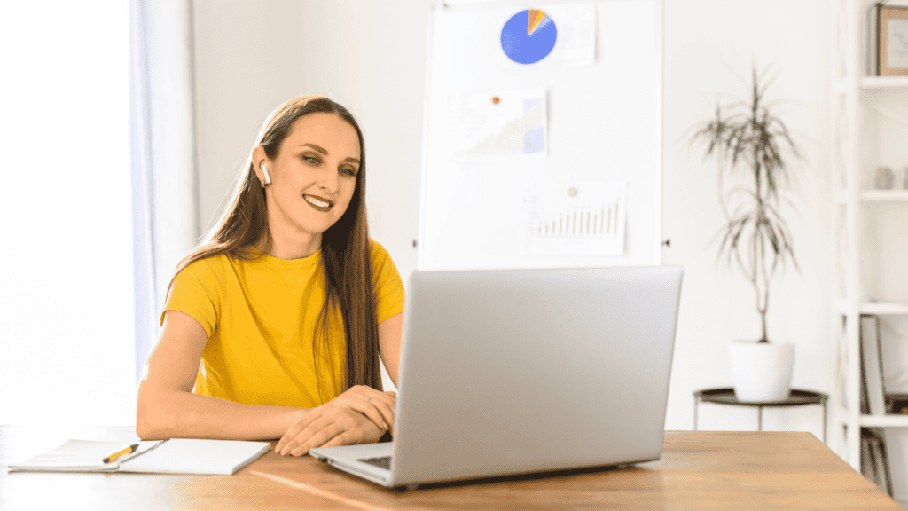 A woman in a yellow shirt working on a laptop, analysing business reports.