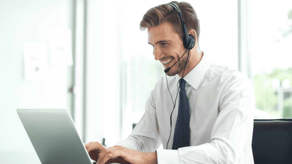 A smiling customer service representative wearing a headset, typing on a laptop in a bright office space.