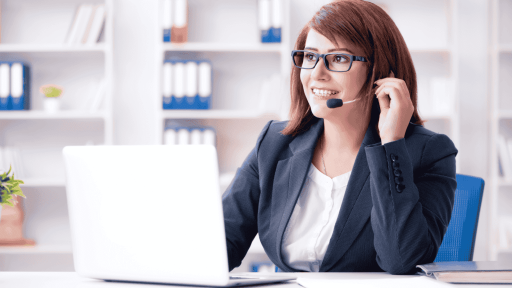 A professional woman wearing glasses and a headset, smiling while working on a laptop in a well-organized office.