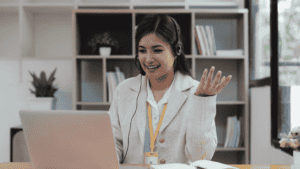 A smiling female customer support representative wearing a headset and a professional white blazer, engaging in a video call with a client.