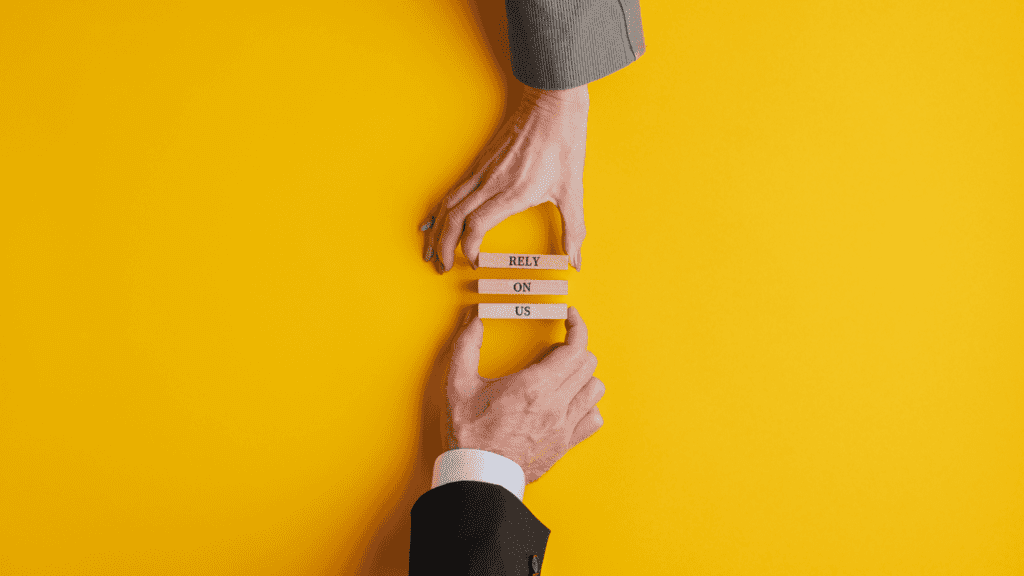 Hands placing wooden blocks with the message "RELY ON US" against a bright yellow background.