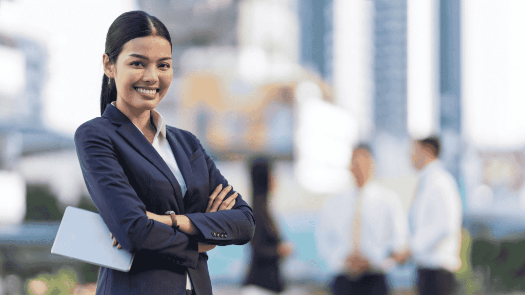 Confident businesswoman in a navy suit holding a tablet with a blurred cityscape and colleagues in the background.