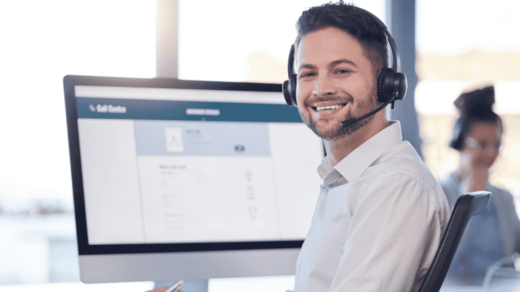 Smiling customer support agent wearing a headset in front of a computer screen displaying a call centre interface.