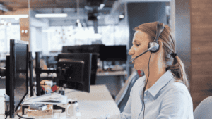 A professional customer support agent wearing a headset, working at a computer in a modern office environment.