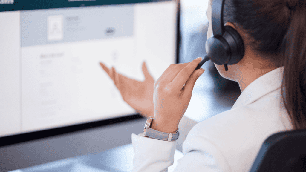 A customer service representative wearing a headset, gesturing towards a computer screen while assisting a customer.