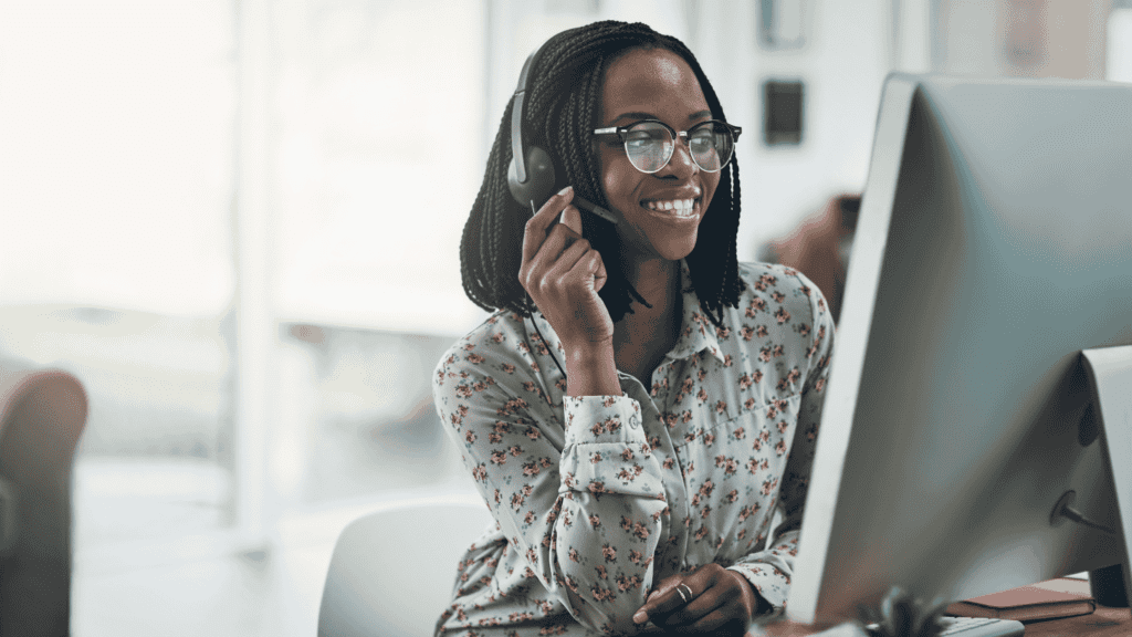 A smiling customer support agent wearing a headset and assisting clients via computer.