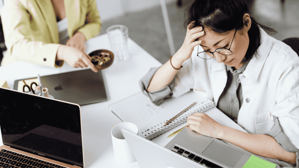 A stressed professional working on data entry tasks with multiple documents and laptops.