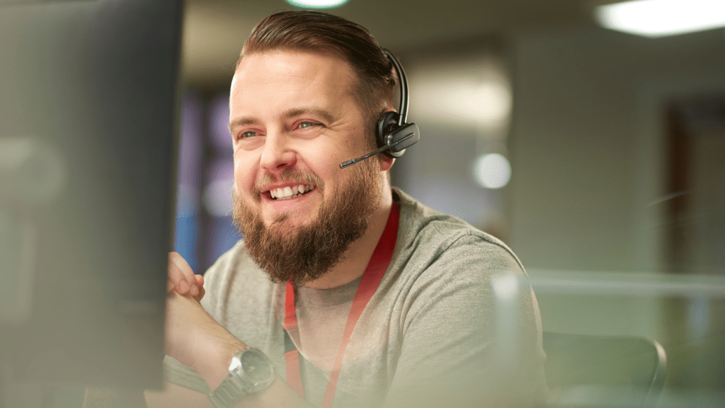 Male customer service representative with a headset, smiling while assisting a customer in a busy support centre.