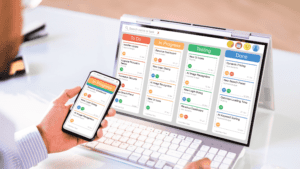 A person using a smartphone and tablet displaying a task management board with "To Do," "In Progress," "Testing," and "Done" columns.