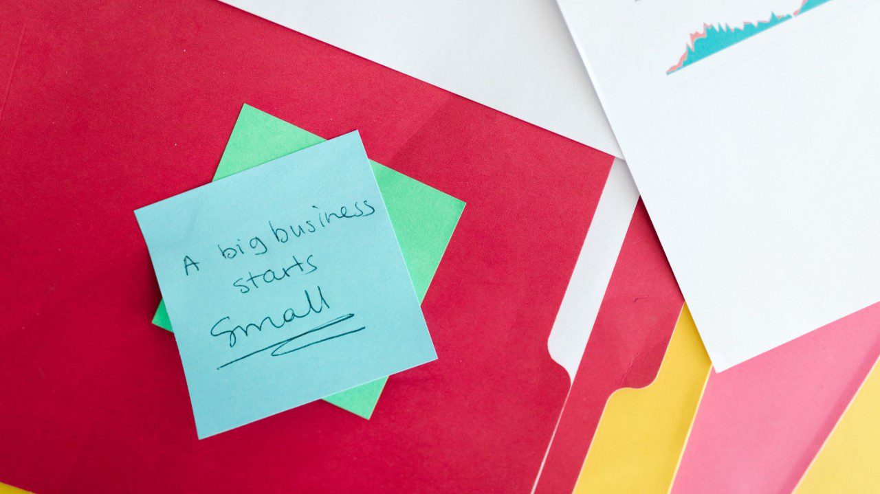 Sticky note reading “A big business starts small” placed on top of colourful folders and documents.