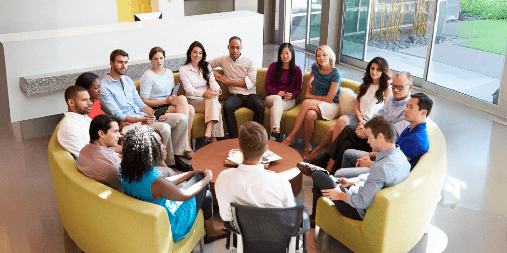 A diverse group of professionals sits in a circle in a modern office, engaged in a relaxed team discussion.