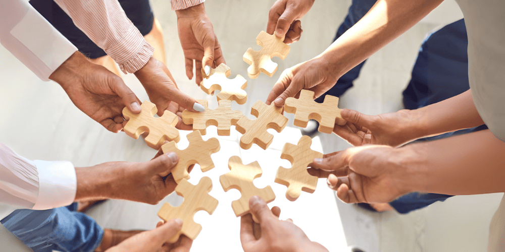 Multiple hands of diverse team members hold large wooden puzzle pieces, working together to fit them into a circle.