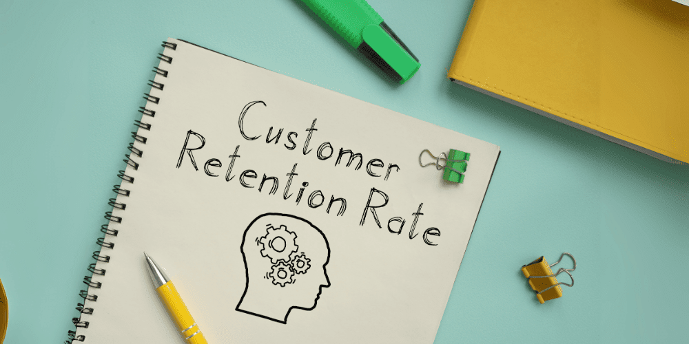 Notebook with the words “Customer Retention Rate” written in bold, next to a drawn outline of a head with gears, on a light green desk.