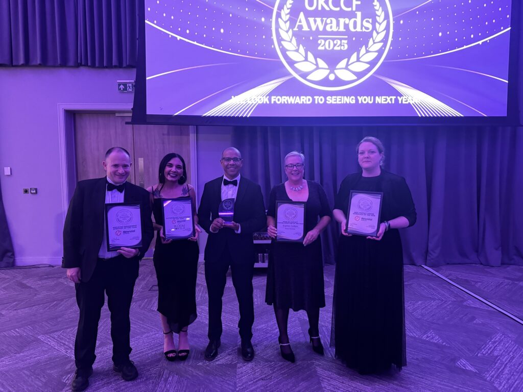Beş Newstel Worldwide ekip üyesi sahnede UKCCF Awards 2025 kupalarını tutarak liderlik, ortaklık ve işyeri kategorilerinde birden fazla kazanımı kutluyor.